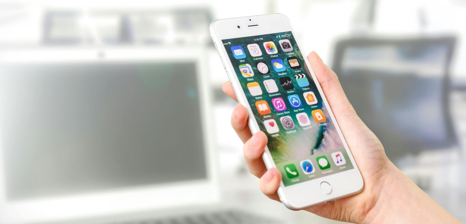 pexels-photo-887751-887751 Close-up view of a smartphone displaying apps, held by a hand, with a blurred laptop in the background.
